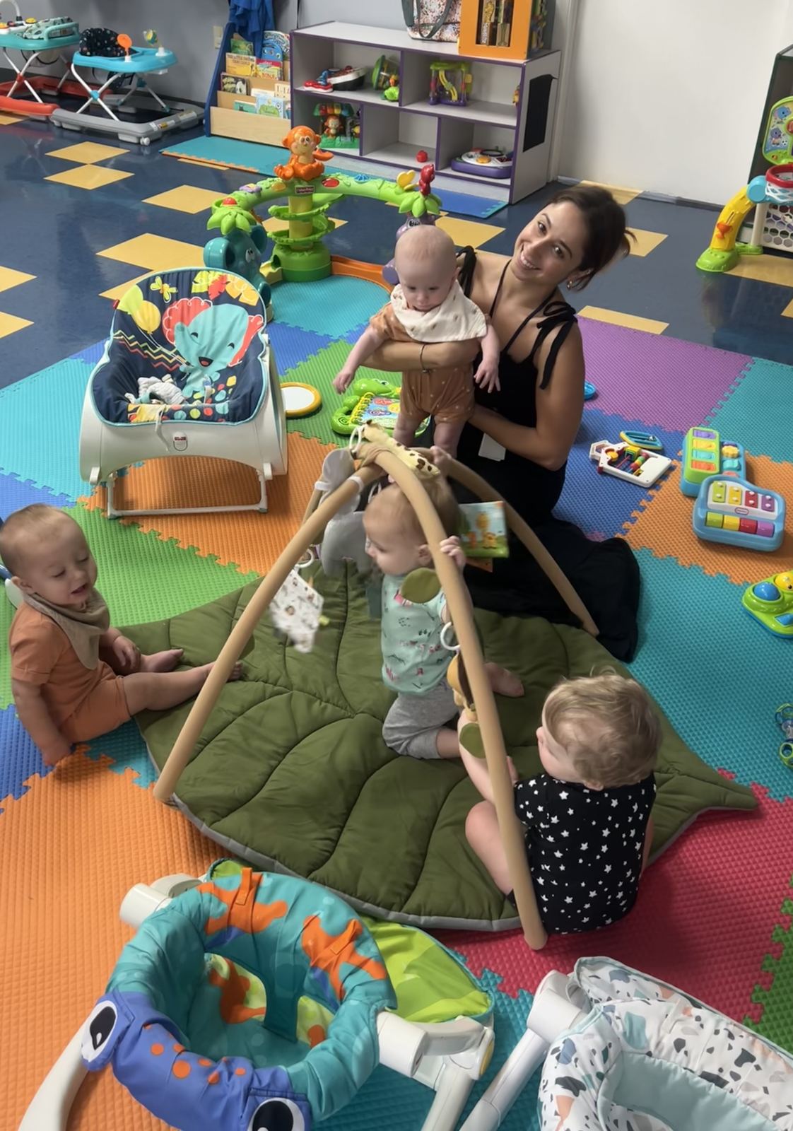 Mrs. Gaby sitting on a play mat, holding a baby while three other infants play nearby
