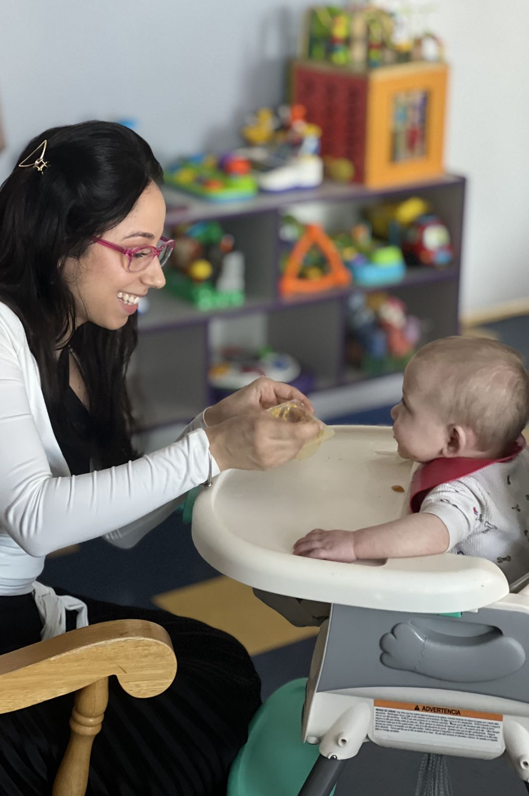 Mrs. Gaby spooning food to a smiling baby seated in a high chair